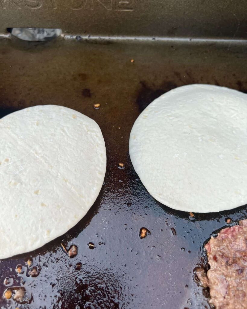 Tortillas on top of a smashed burger on a griddle. 