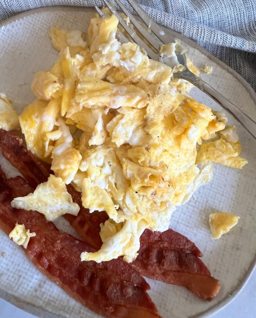 Scrambled Eggs next to bacon on a plate with a fork. 